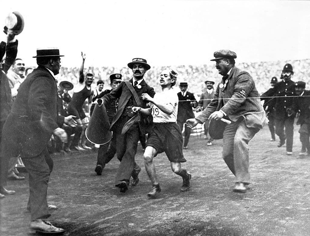 Black-and-white photo of Dorando Pietri, exhausted, being supported by officials as he crosses the finish line in the 1908 London stadium