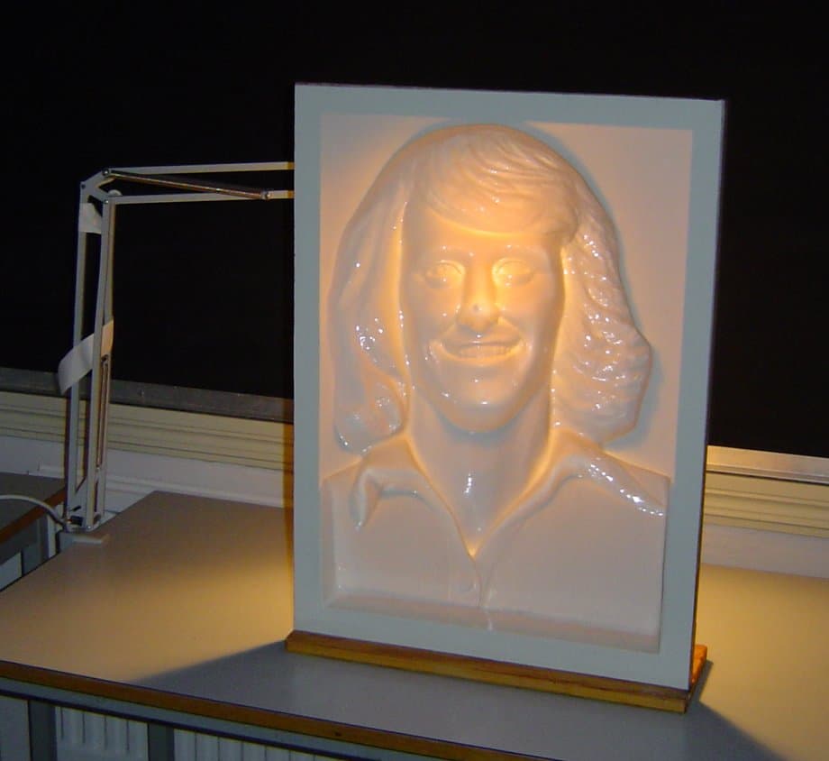 A white mask of a smiling face mounted in a frame and lit by a desk lamp; it looks like a face bulging outward.