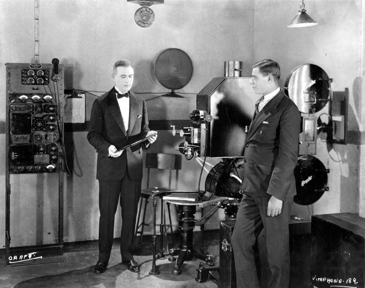 A 1926 black-and-white photograph of two men in a projection room beside a large Vitaphone film projector fitted with a record turntable.