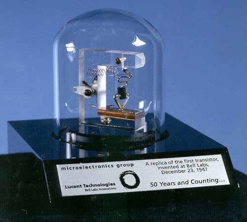 A replica of the first point-contact transistor: a small triangular plastic wedge with two gold wires pressed onto a germanium crystal, mounted on a metal base.