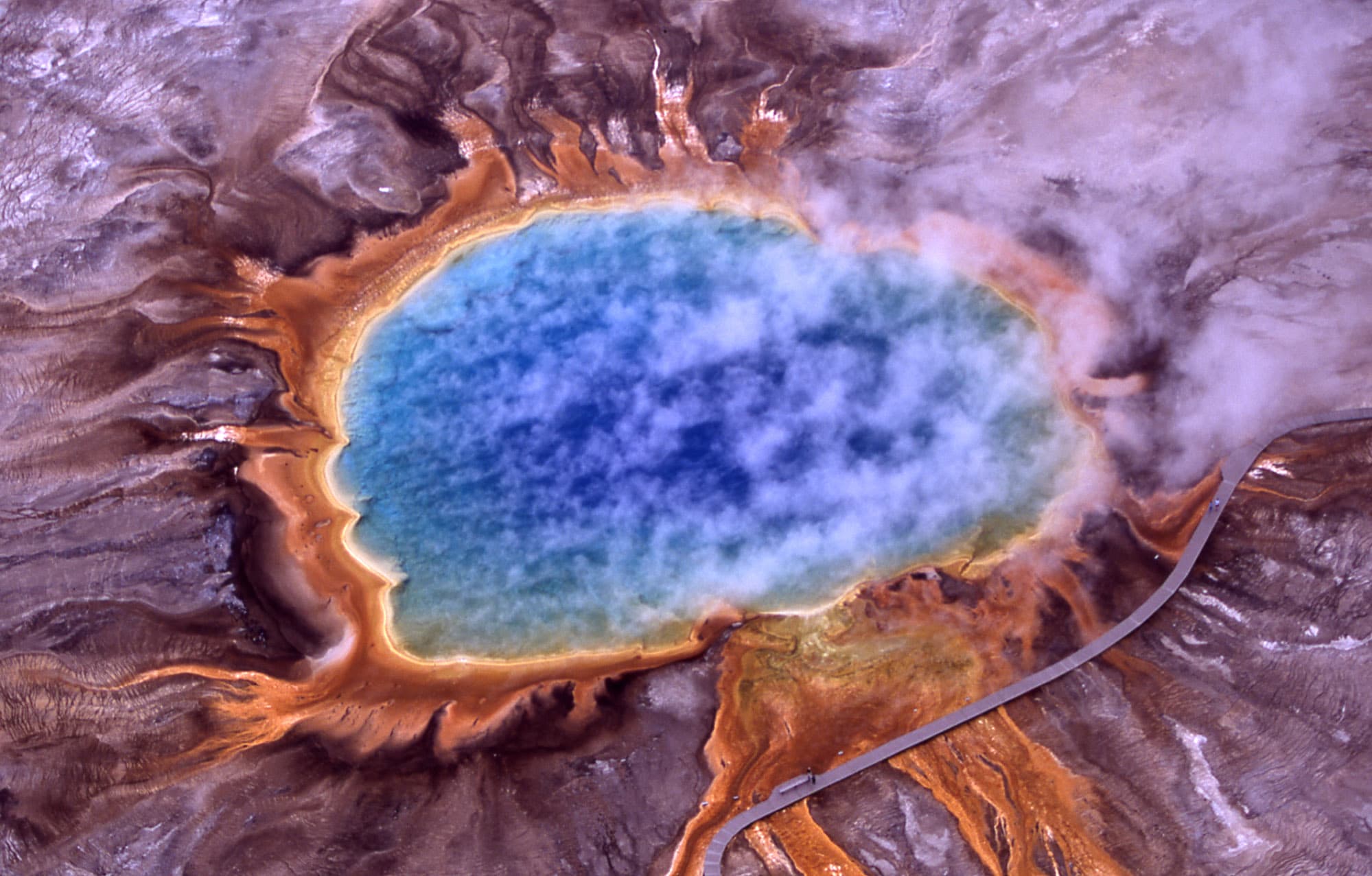 Aerial view of a large, brightly colored hot spring in Yellowstone National Park, with rings of orange, yellow, green and blue around a deep blue center.
