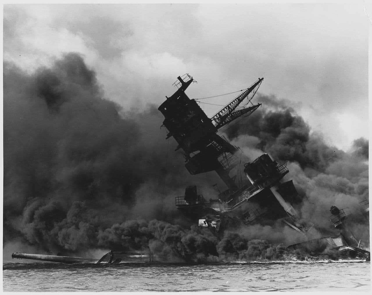Black-and-white photo of the battleship USS Arizona burning and billowing thick smoke at Pearl Harbor after the Japanese attack.