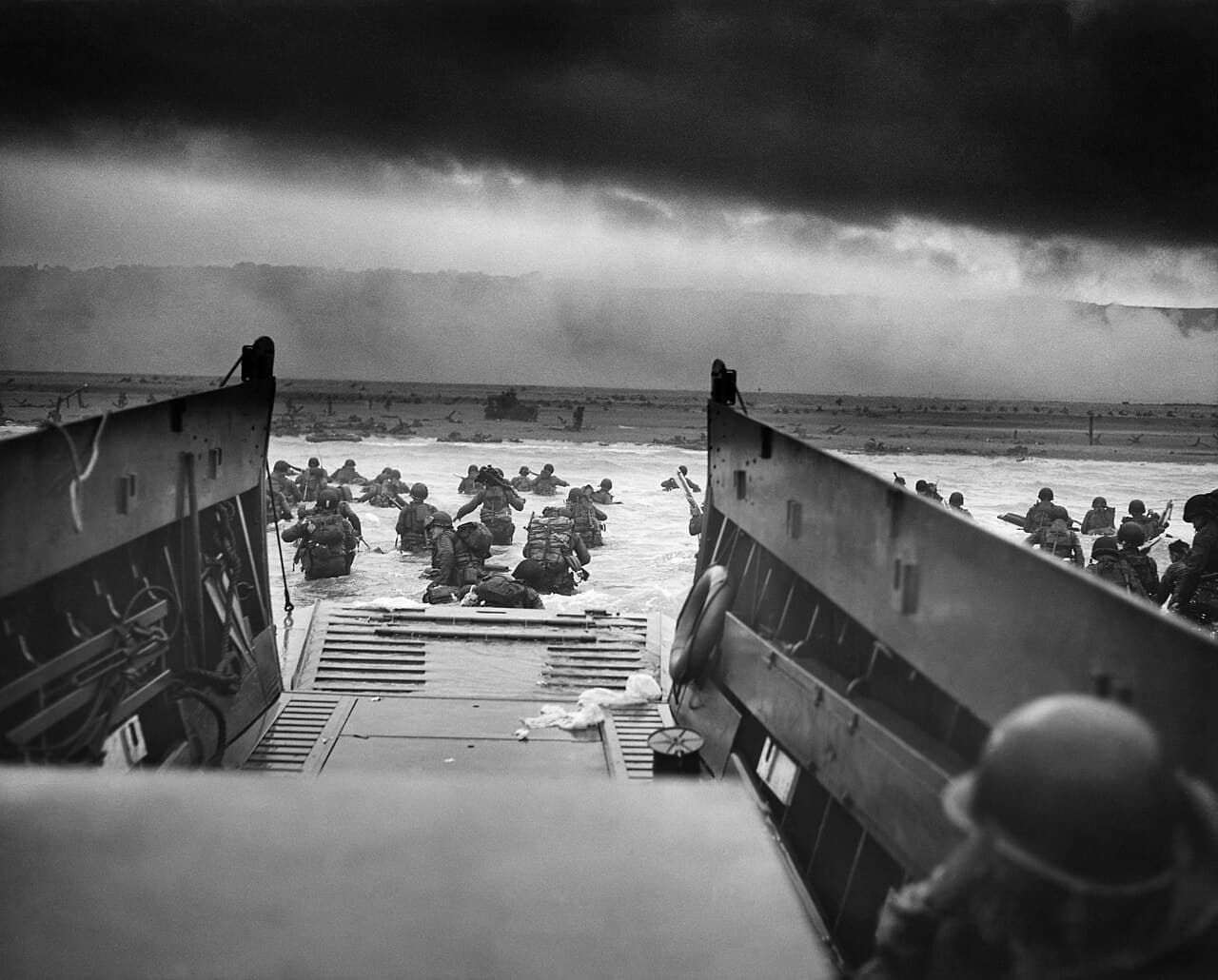 Black-and-white photo of American soldiers wading off a landing craft into waist-deep water under fire at Omaha Beach.