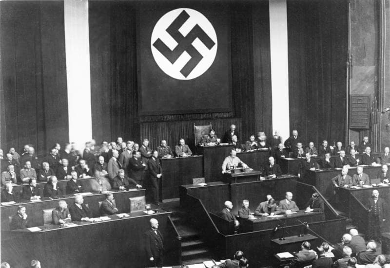 Black-and-white photo of Hitler speaking at a lectern in the Kroll Opera House, with deputies and a large eagle emblem behind him.