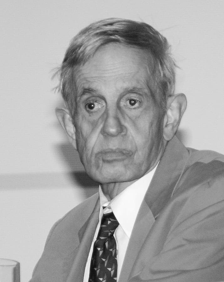 John Nash, an elderly man with white hair and glasses, speaking at a game theory symposium.