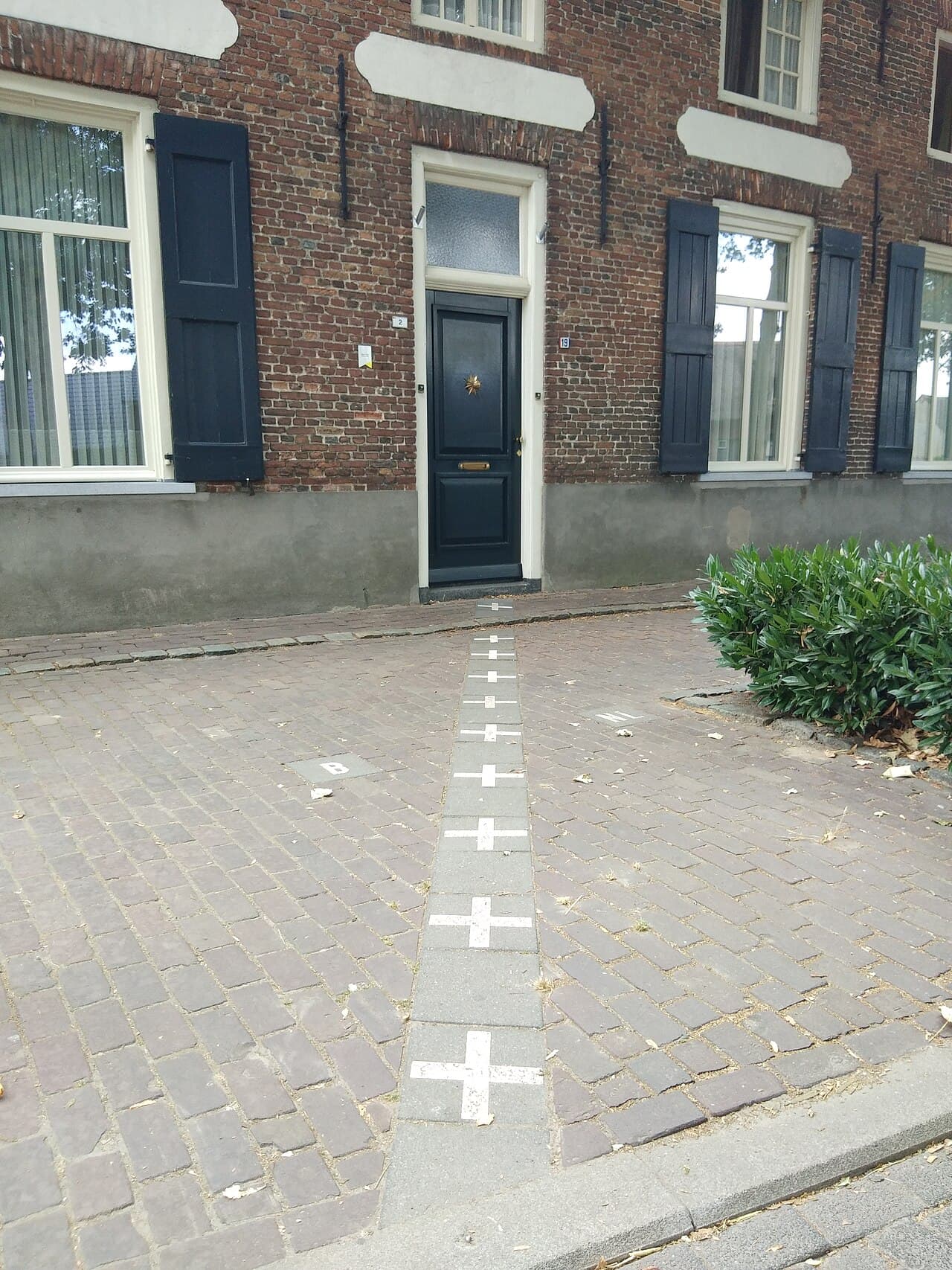 A brick house with a dark front door; a line of white crosses set into the cobbled pavement runs straight up to the doorway.