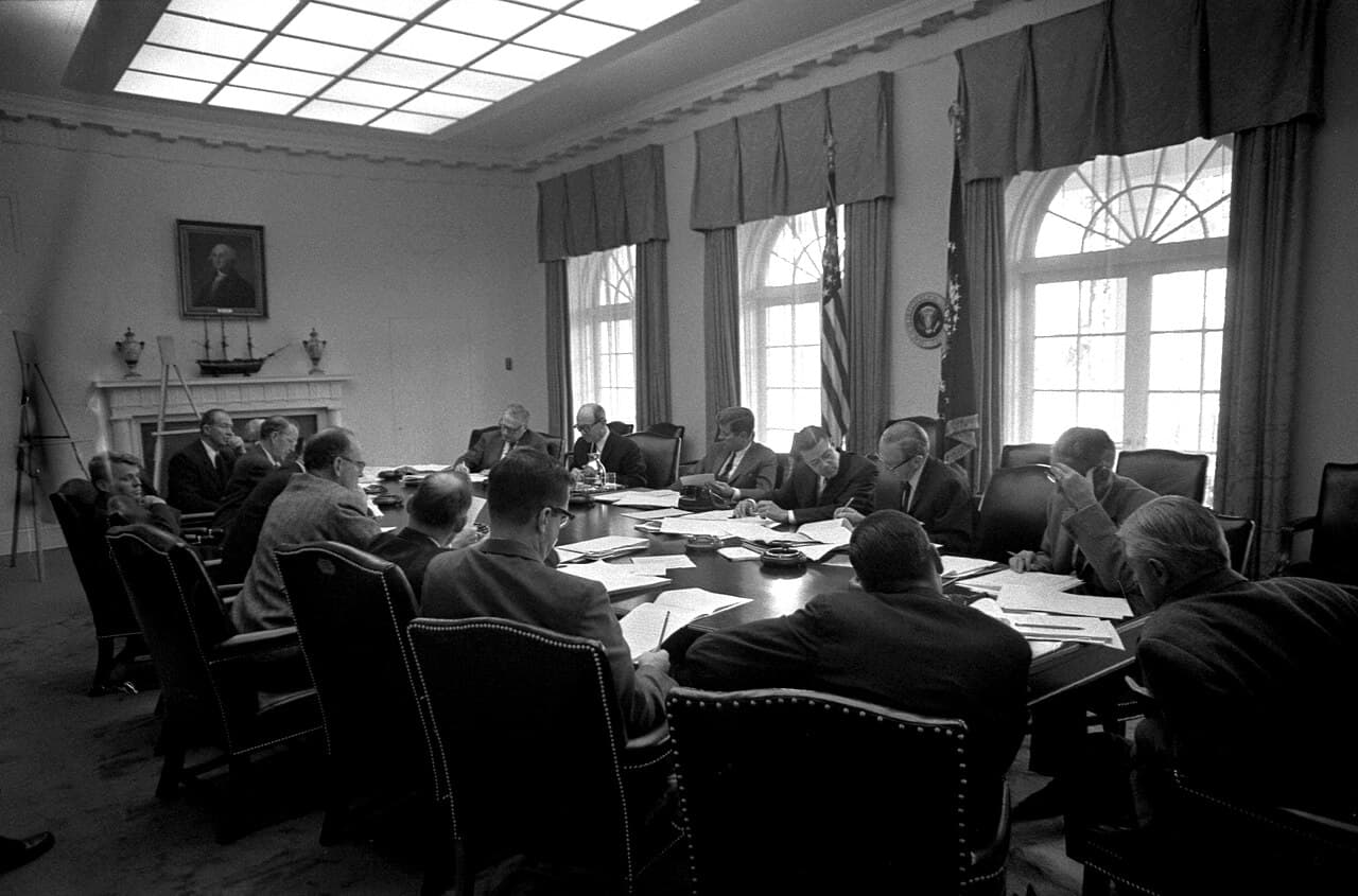 Black-and-white photograph of President Kennedy and his advisers seated around a table during an EXCOMM meeting in the White House.