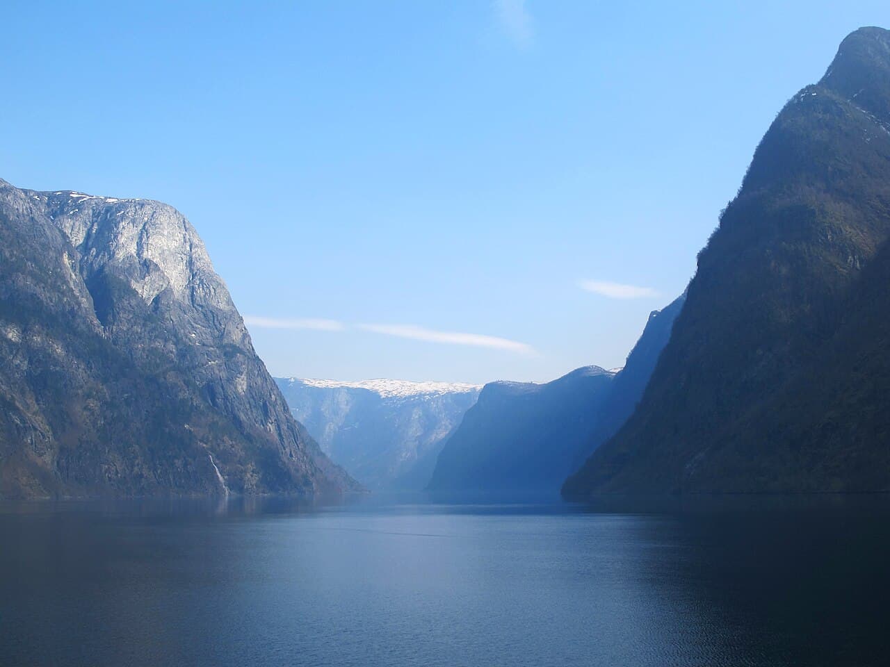 A long, narrow Norwegian fjord with steep, snow-dusted mountains rising directly from the dark water on both sides.