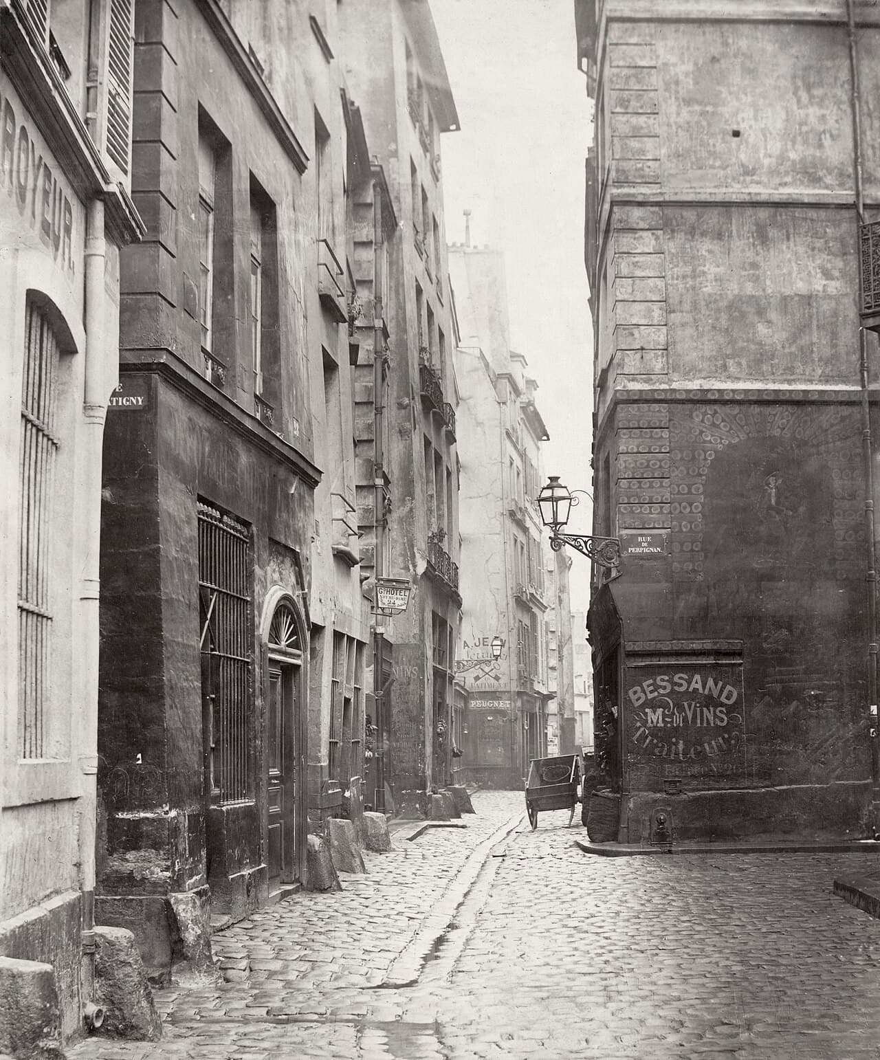 Old black-and-white photograph of a narrow cobbled Paris street with a gutter running down the middle and tall, uneven houses on both sides.