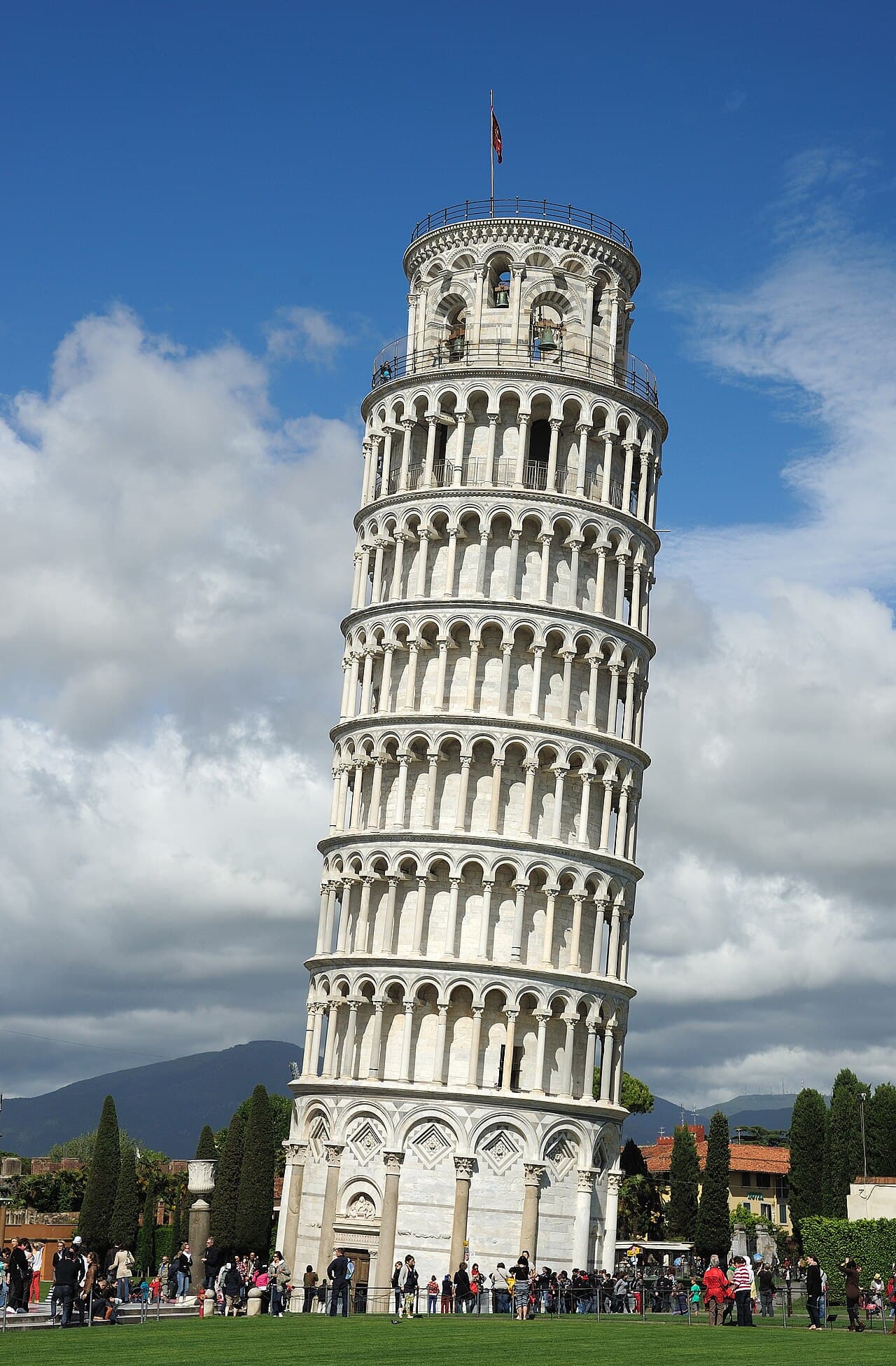 The white marble Leaning Tower of Pisa, a round bell tower with stacked arcades, visibly tilted to one side against a blue sky.