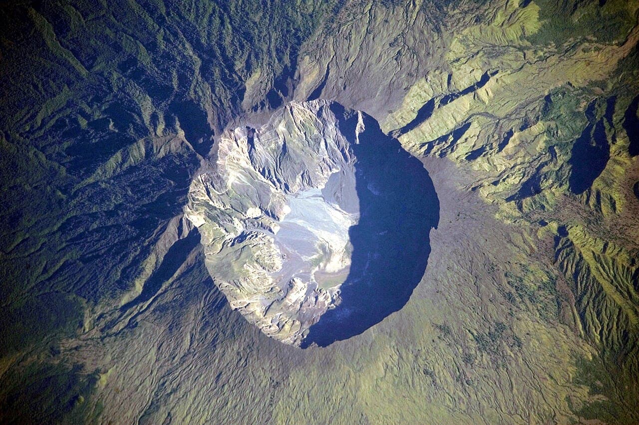 Astronaut photograph of Mount Tambora seen from above: a vast, deep crater surrounded by green slopes.