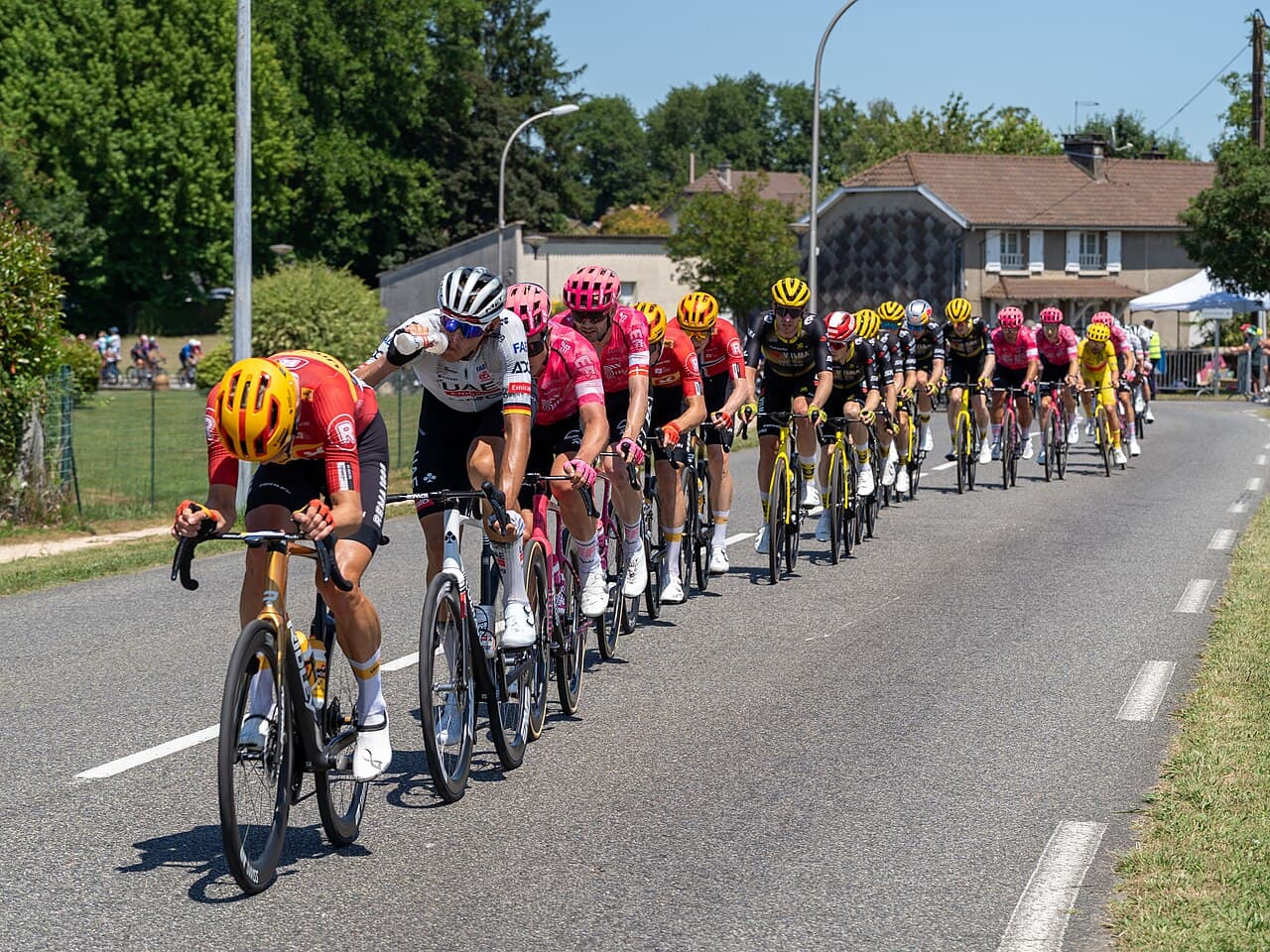 A dense pack of racing cyclists in colourful team jerseys riding close together along a road during the Tour de France.